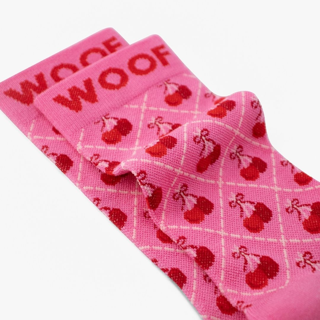 A Cherry Bows Bamboo Crew Compression Socks folded neatly to highlight the dark pink woven "WOOF" cuff bands and the cherry-and-bow lattice knit pattern layout.  