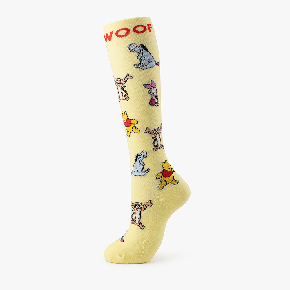 Studio shot of the pale yellow pair from the Disney’s Winnie the Pooh Bamboo Compression Socks 4 Pack, highlighting a red "WOOF" cuff band and full-body illustrations of Pooh, Tigger, Eeyore, and Piglet.   